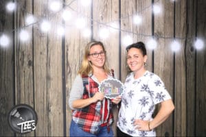 Two friends posing with sign at rustic backdrop framed by warm string lights