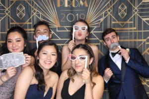 Friends in formal attire holding props in front of prom 2019 art deco backdrop