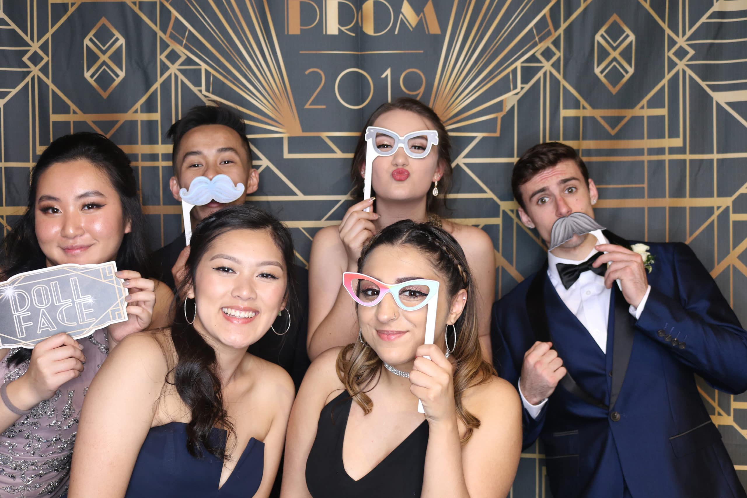 Friends in formal attire holding props in front of prom 2019 art deco backdrop