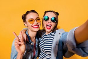 Two young women taking phone selfie with sunglasses on yellow background