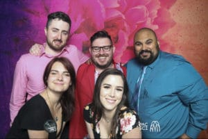 Five friends posing closely together in front of pink watercolor backdrop