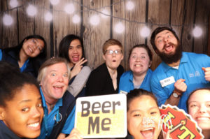 Group of coworkers holding Beer Me sign at rustic string-lit photo booth