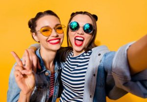 Two friends taking smiling selfie wearing sunglasses on yellow background