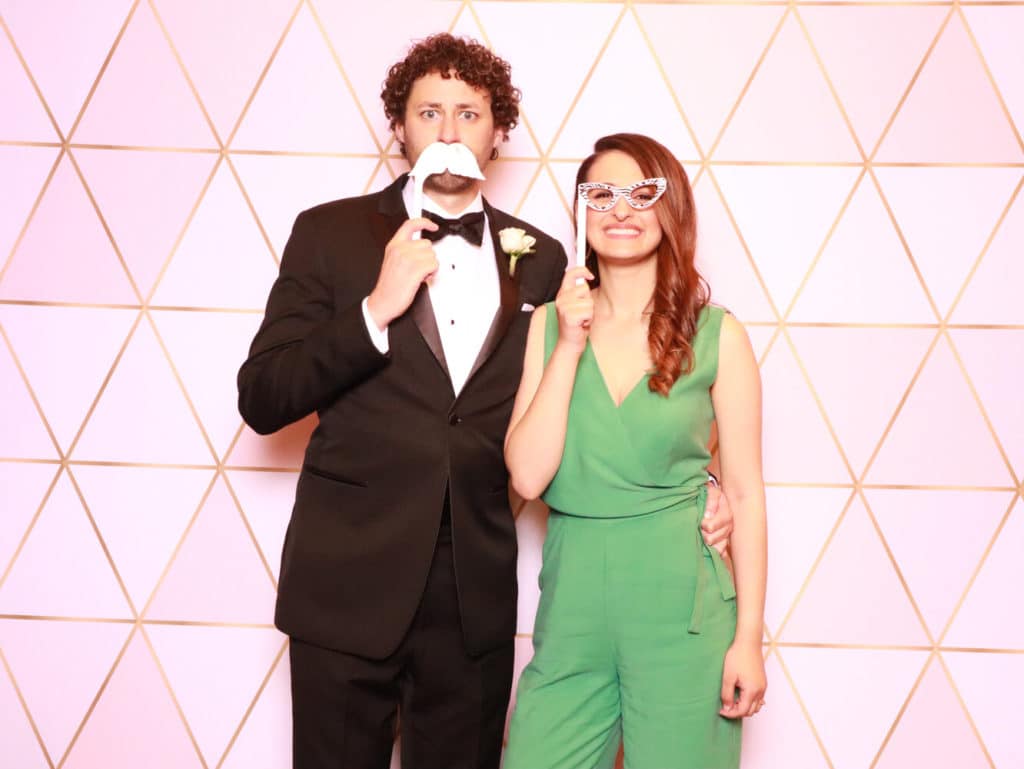 Couple holding mustache and glasses props at pink geometric photo booth backdrop