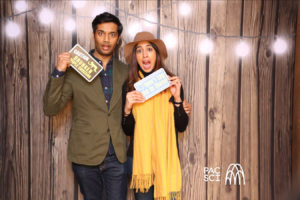 Couple holding ticket and message props at rustic string-lit photo booth