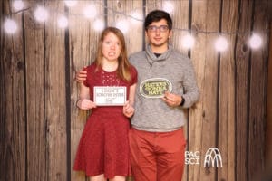 Couple holding sign props at rustic wood photo booth with string lights