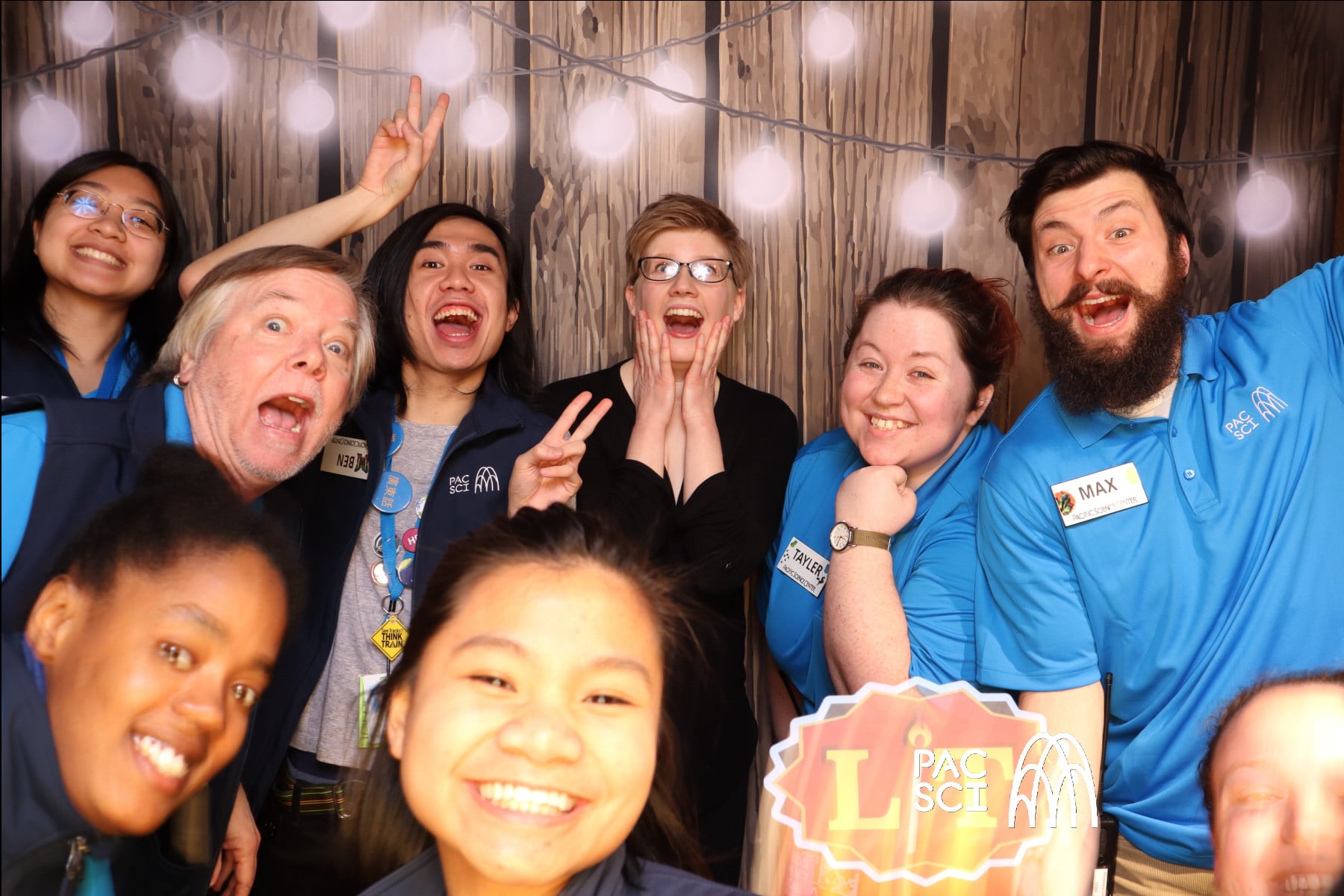 Group of coworkers crowded together making faces at rustic string-lit photo booth