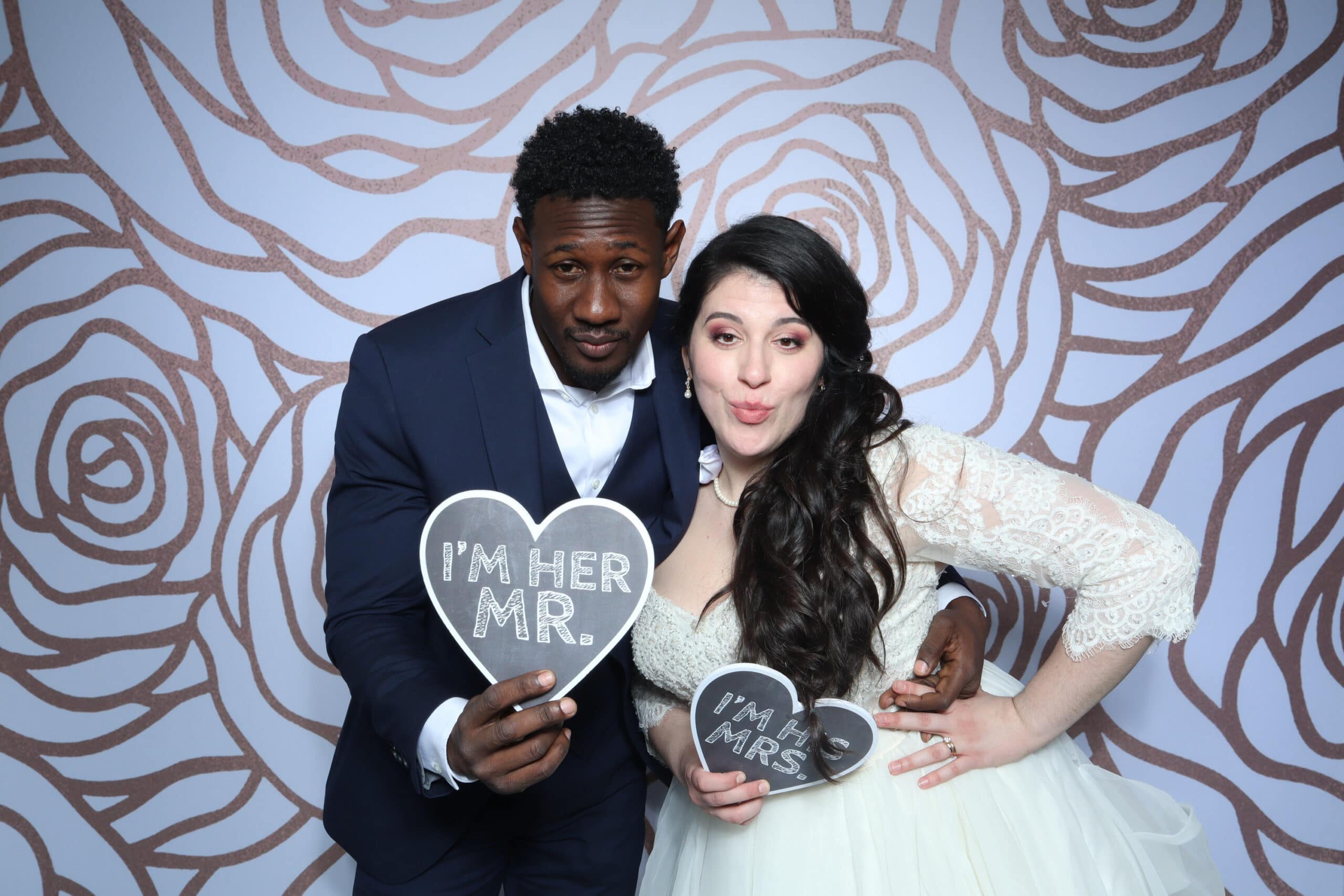 Couple holding I'm Her Mr and I'm His Mrs heart props at floral wedding backdrop