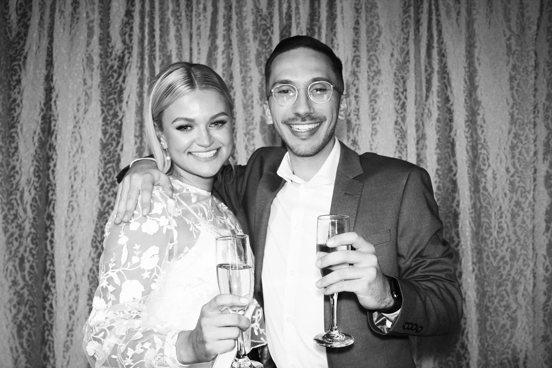 Black and white photo of couple holding champagne flutes at lace-curtain photo booth