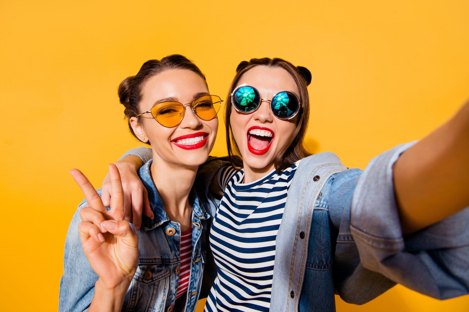 Two young women in sunglasses taking smiling selfie on yellow background