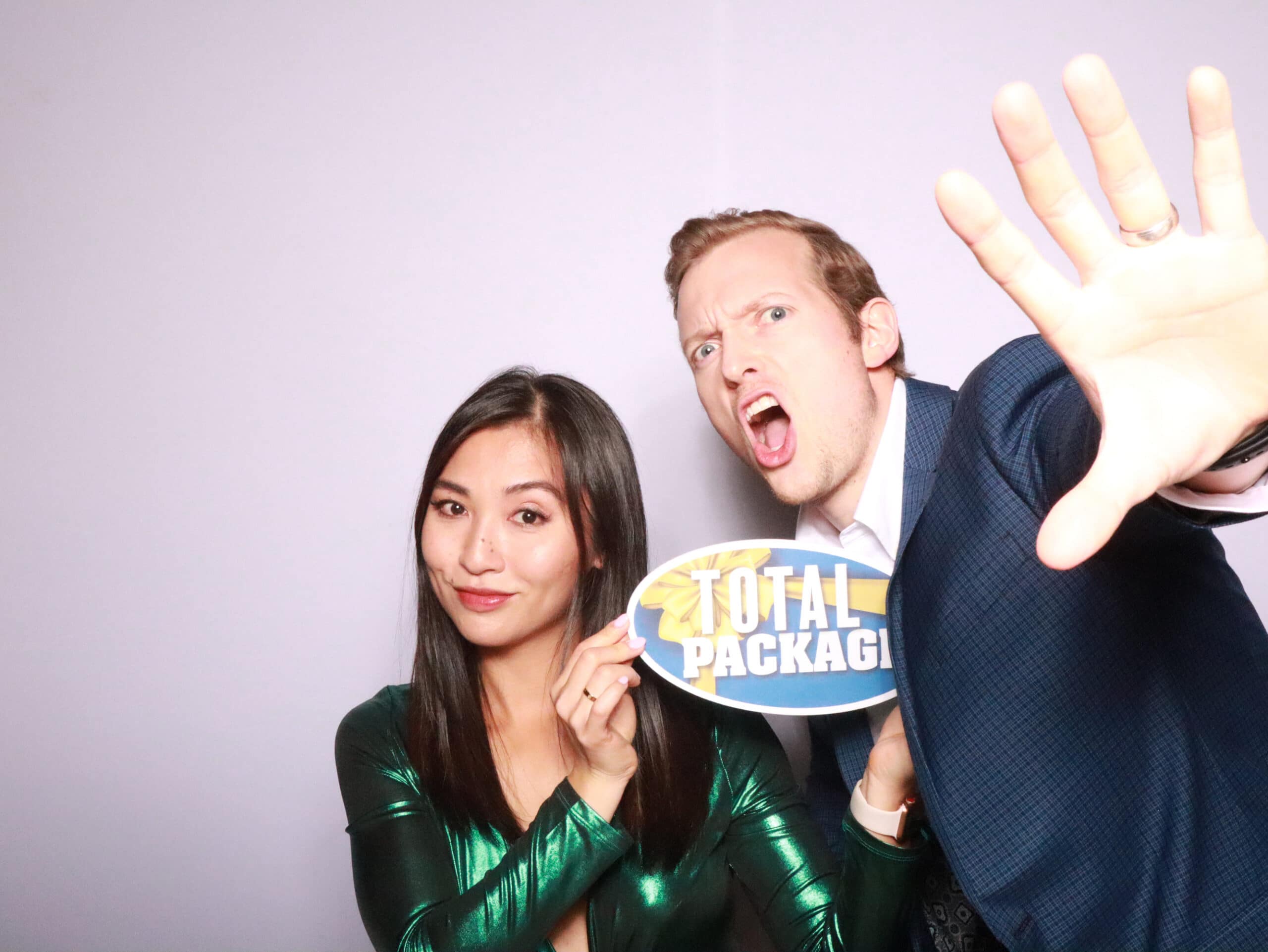 Couple holding Total Package sign making playful faces at photo booth on lavender backdrop