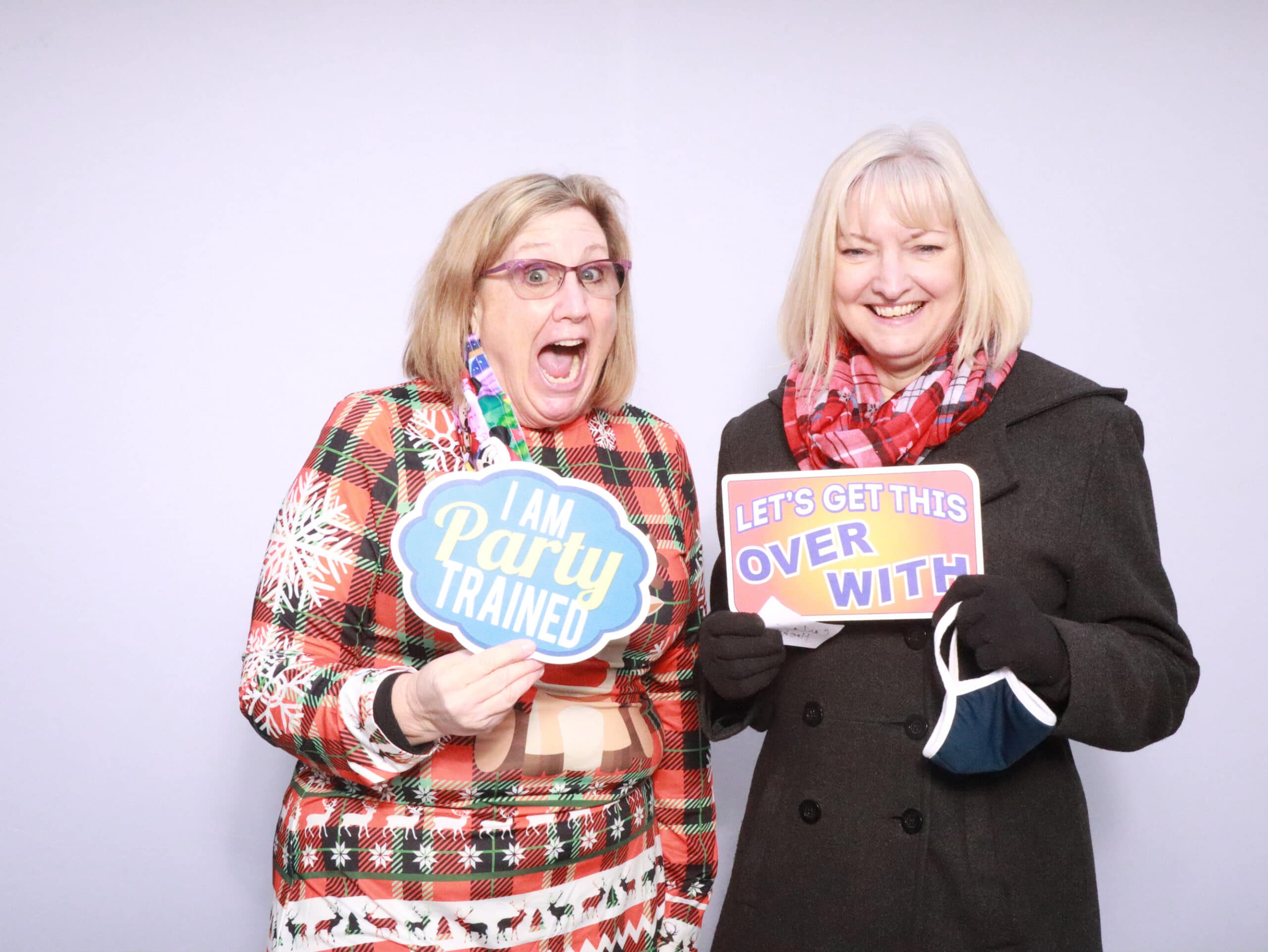 Two women in holiday sweaters holding playful sign props at photo booth