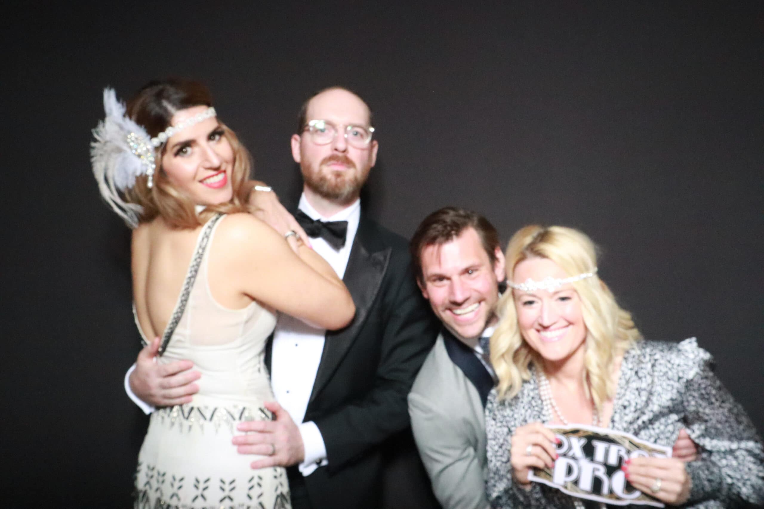 Two couples in formal attire posing playfully at black photo booth backdrop