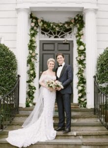 Bride and groom posing at white doorway with greenery garland and bouquet