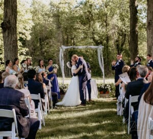 Bride and groom kissing at outdoor woodland wedding ceremony with applauding guests