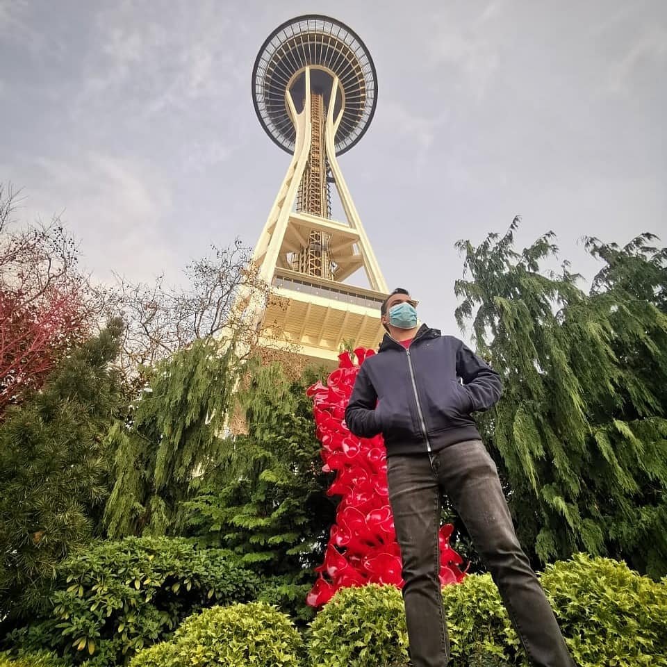 Person in mask standing in front of Seattle Space Needle with red Chihuly glass