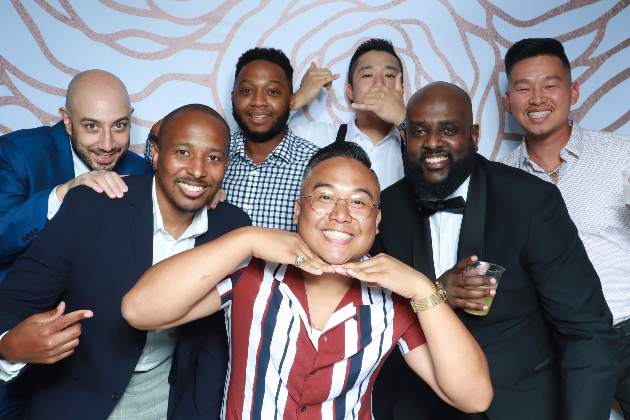 Group of men in formal attire posing playfully at white photo booth backdrop