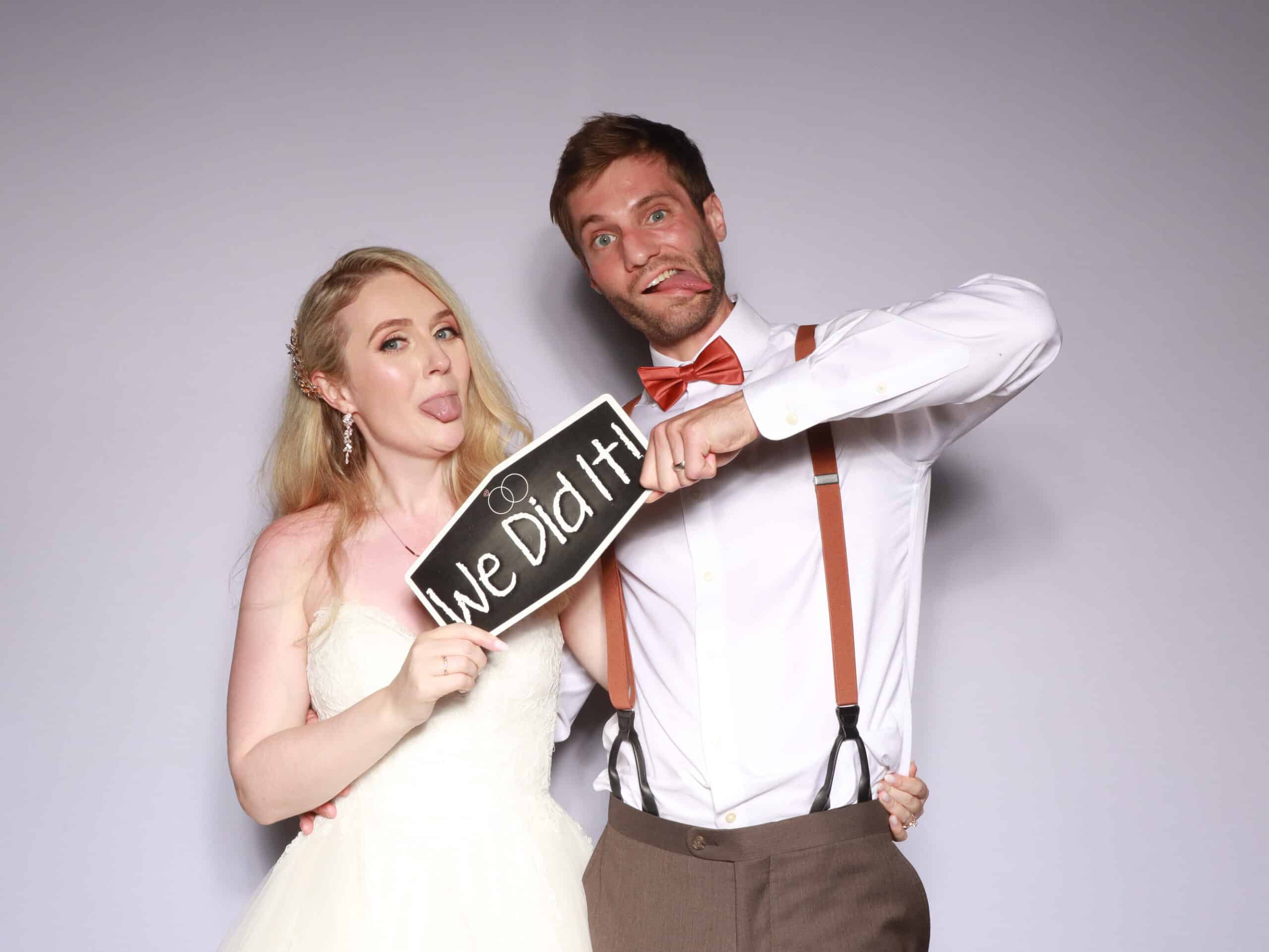 Bride and groom holding We Did It sign at photo booth with white backdrop