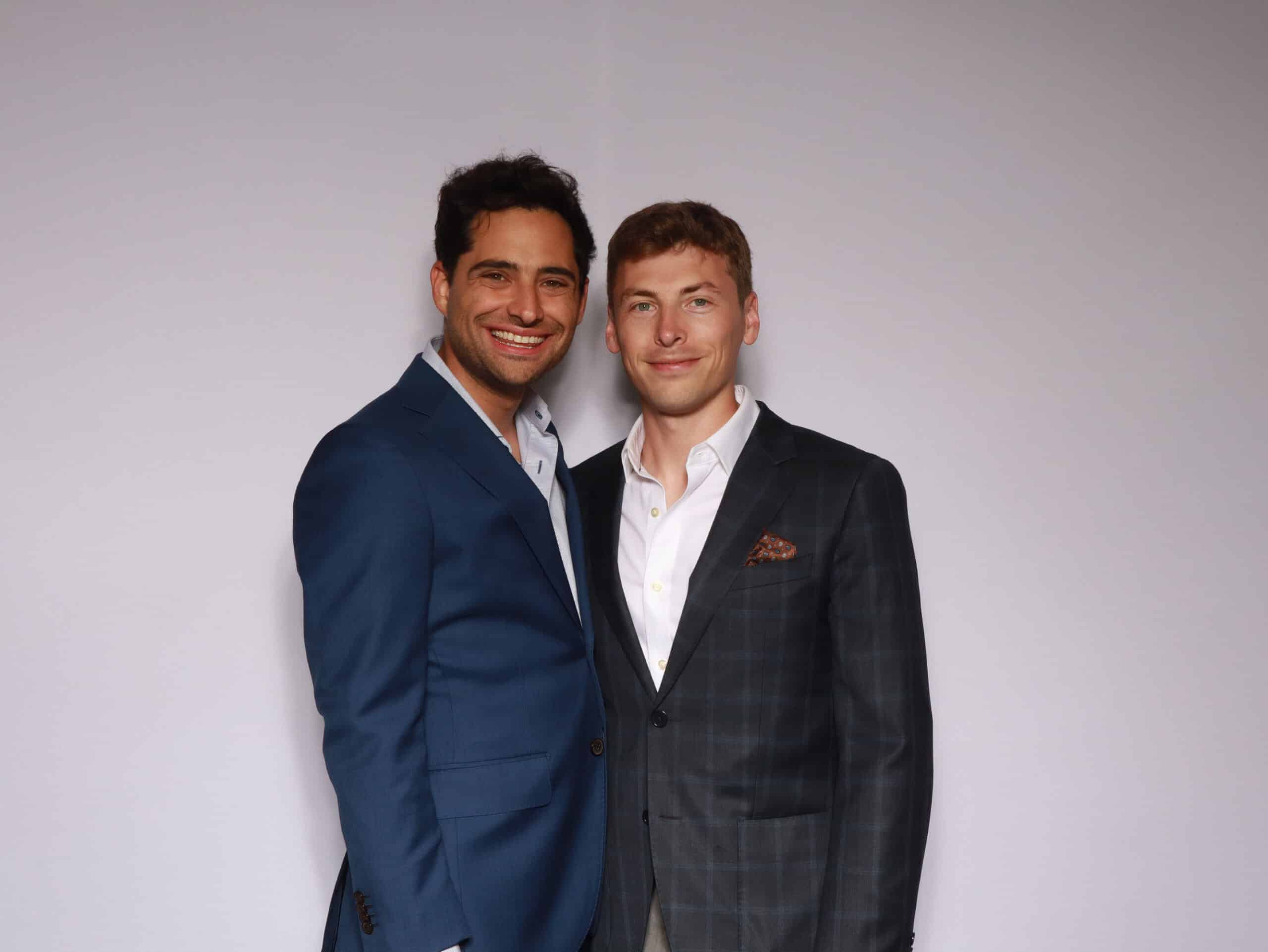 Two men in formal suits posing closely together at white photo booth backdrop