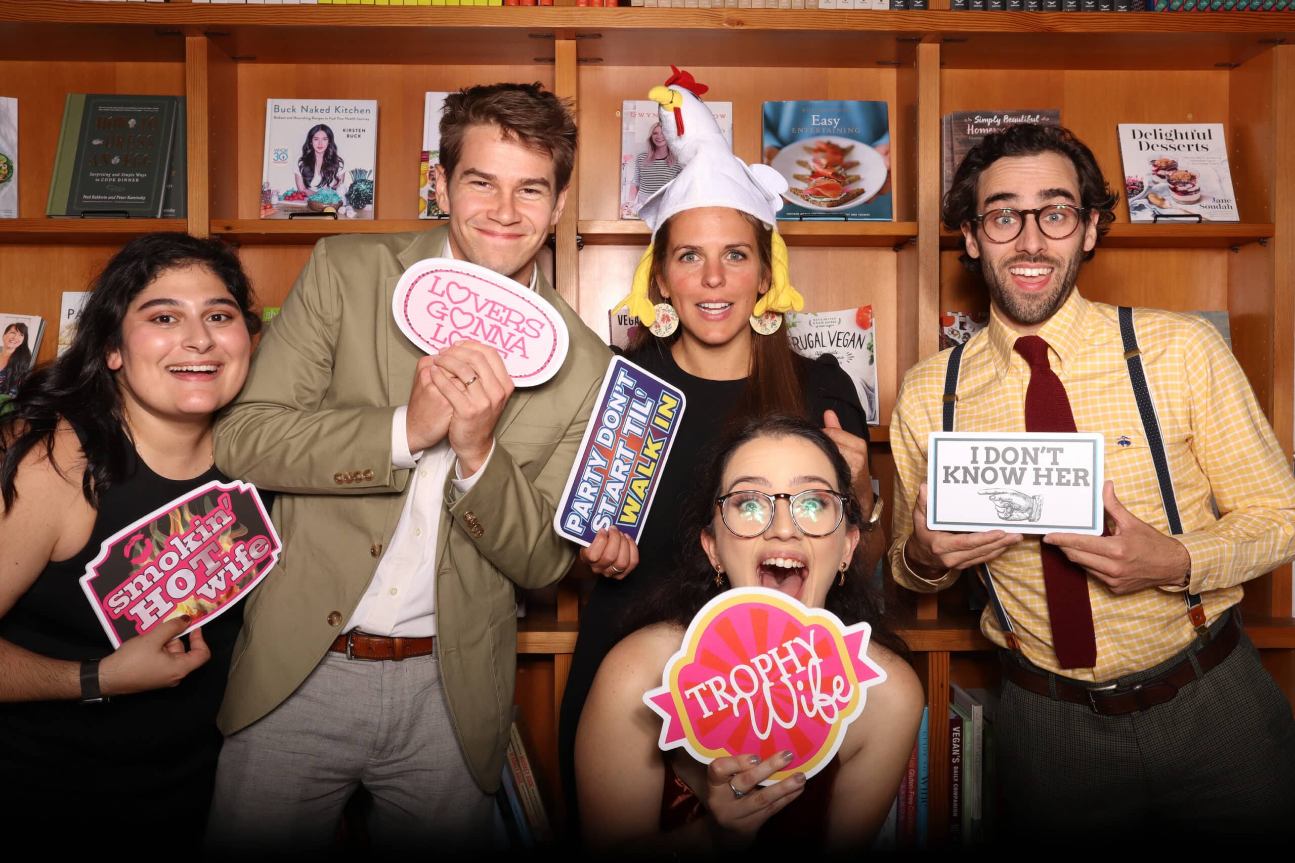 Group holding Trophy Wife and I Don't Know Her sign props at bookshelf backdrop