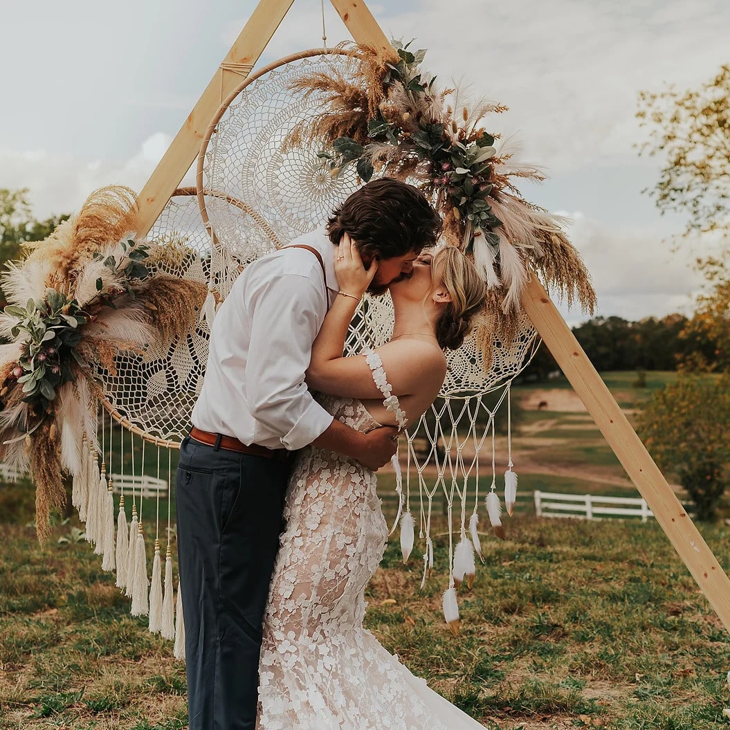 Bride and groom kissing at boho outdoor wedding with pampas grass triangle arch