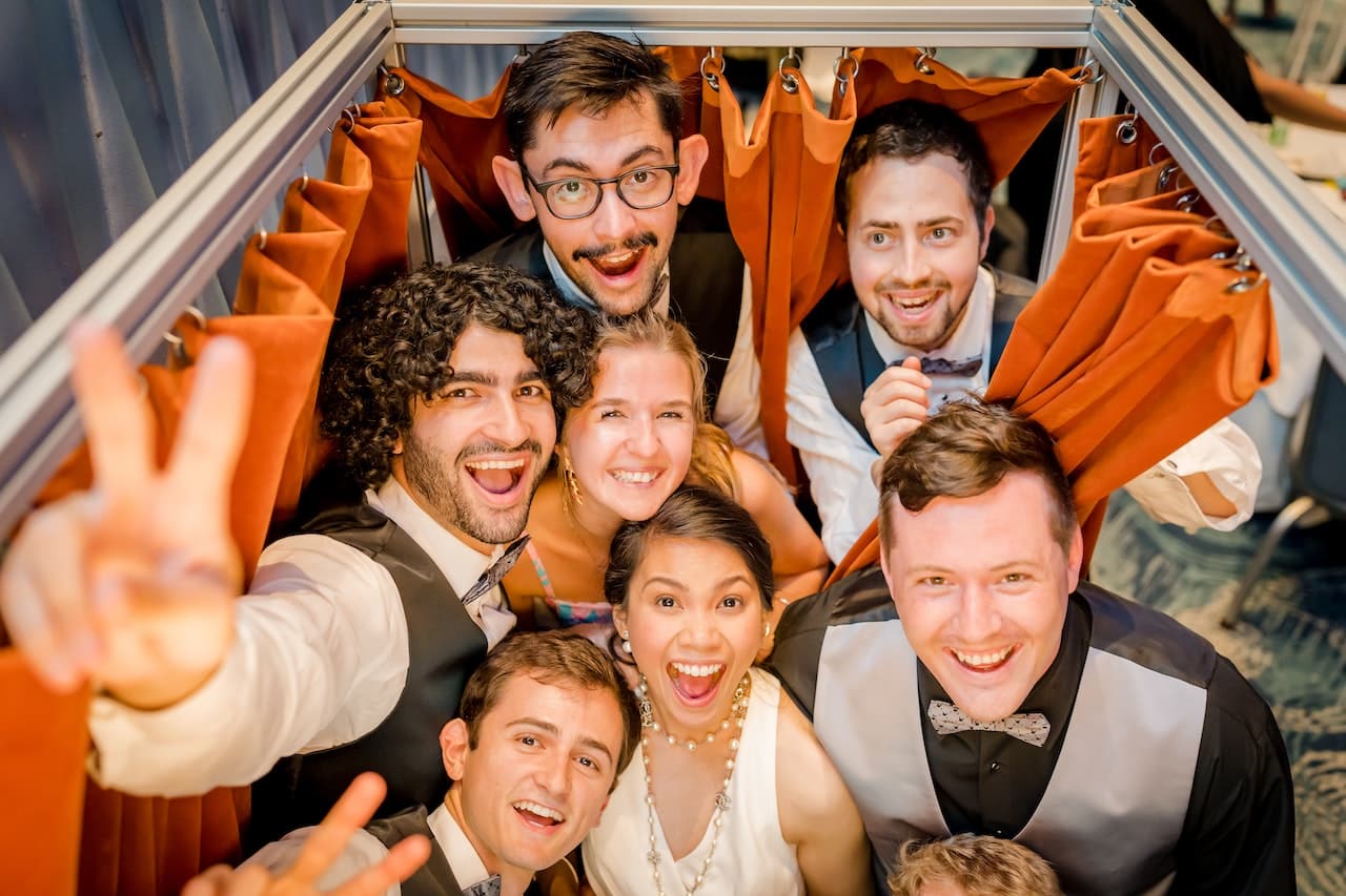 Group of friends in formal attire making playful peace signs at orange-curtained booth