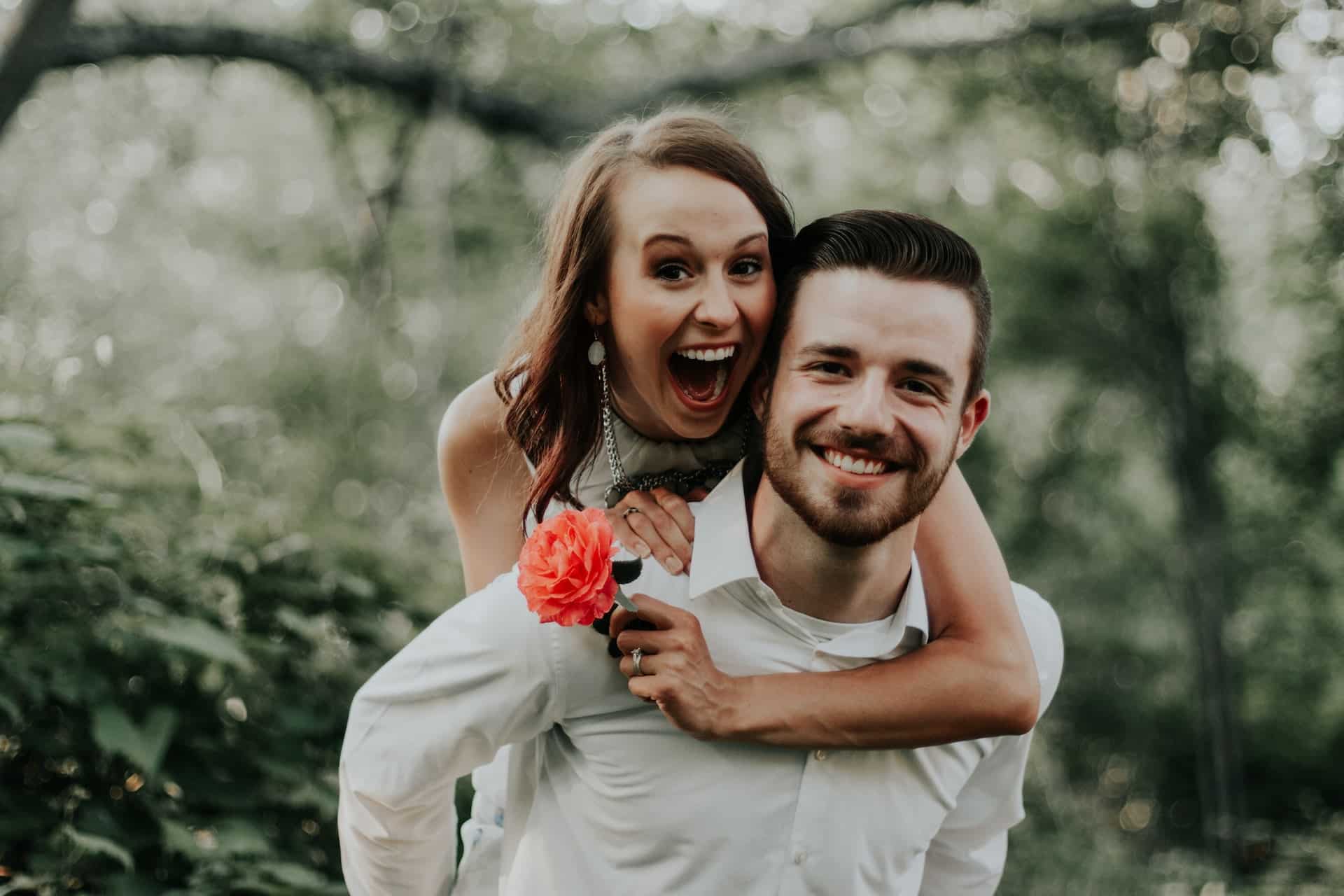 Couple piggyback laughing in lush green outdoor setting with flower