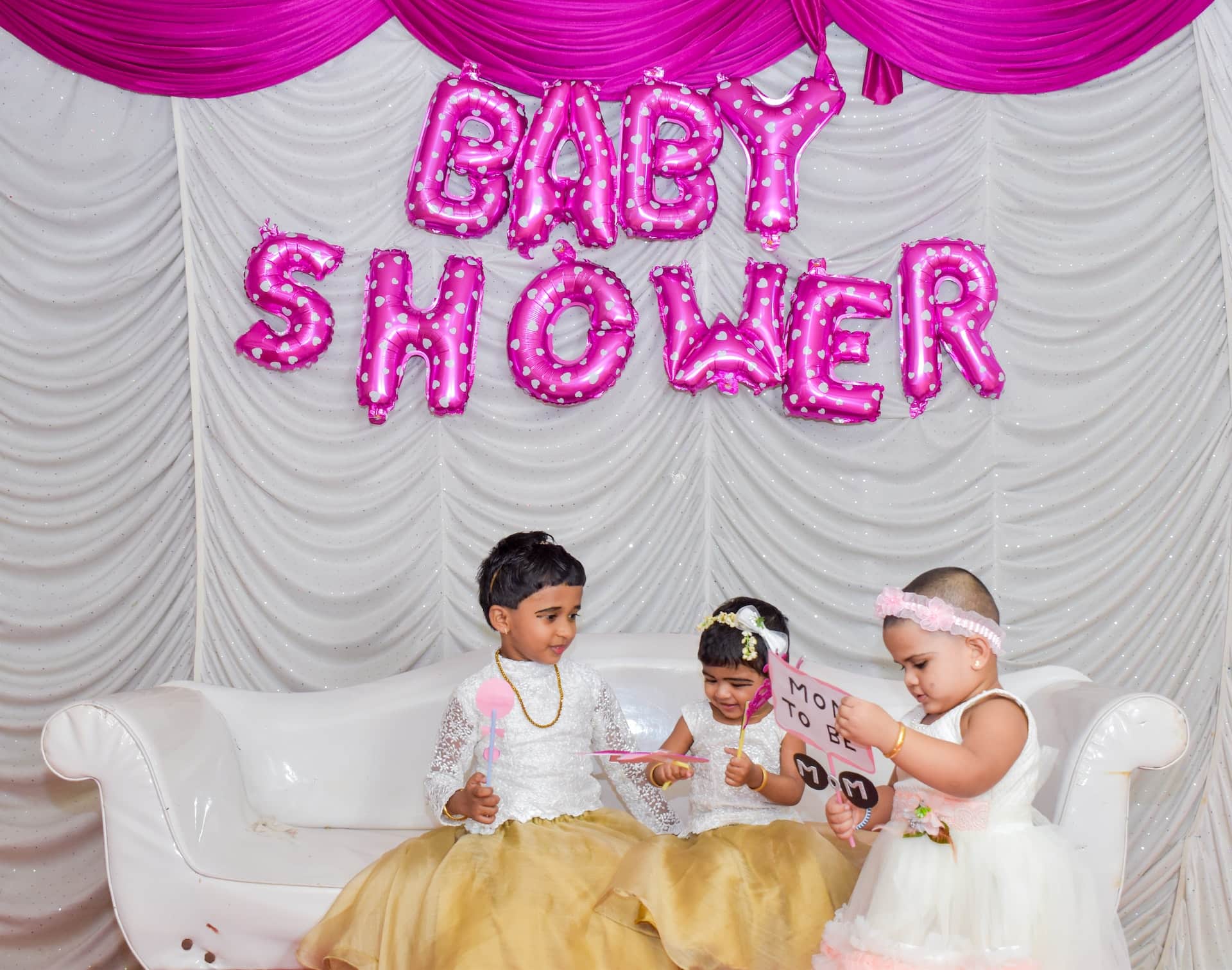 Three young children on couch at pink Baby Shower balloon backdrop