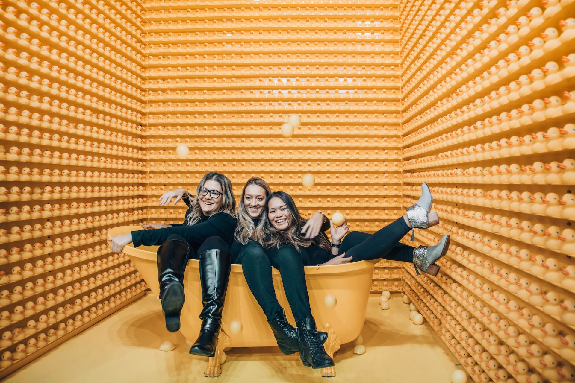 Three women laughing together inside artistic yellow ball pit installation