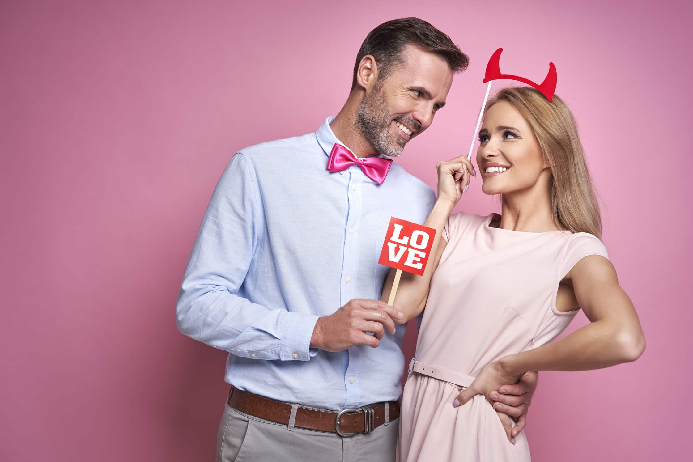 Couple posing with Love sign and devil horns at pink Valentine's Day backdrop