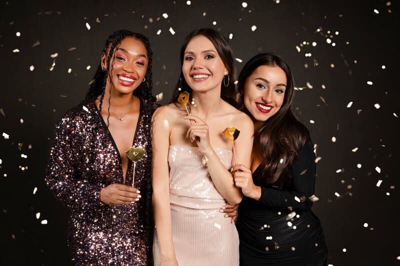 Three women in formal attire celebrating with confetti and props at black backdrop