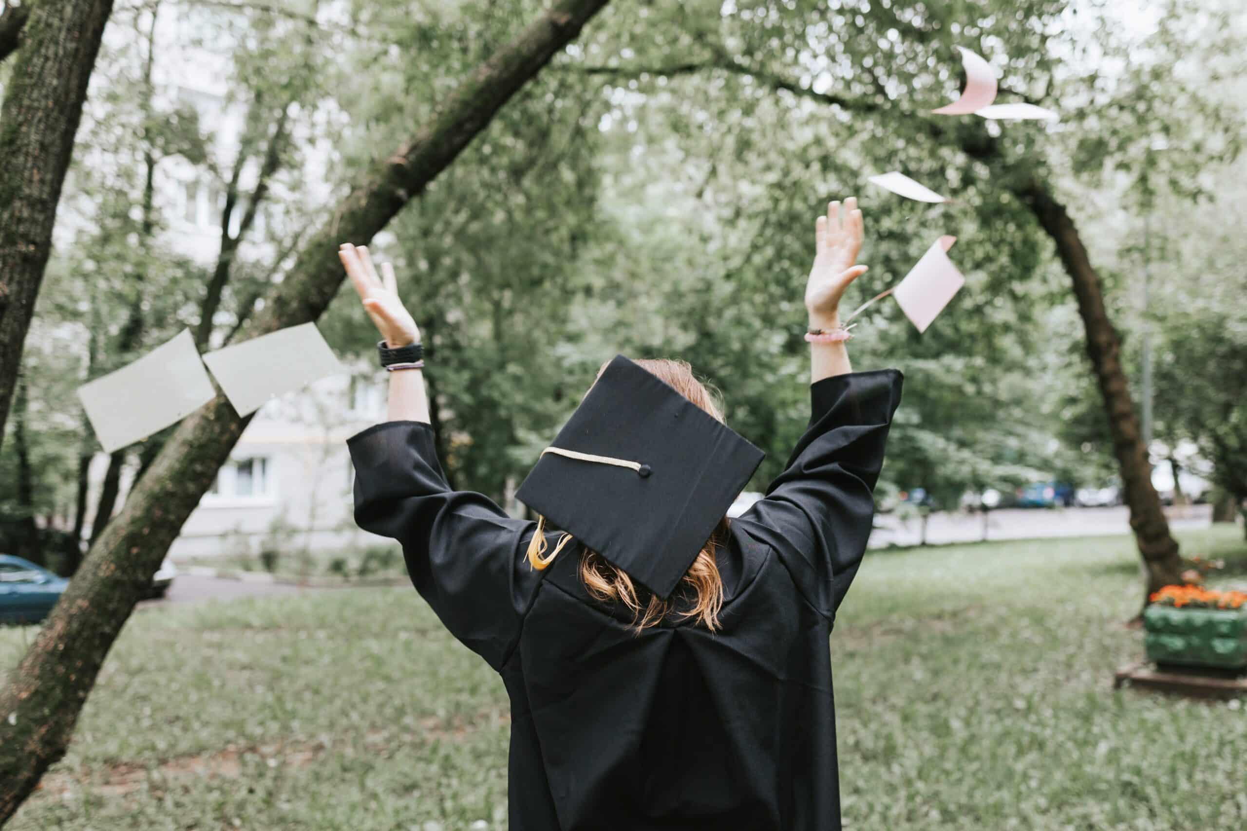 Graduate in cap and gown throwing diploma into the air outdoors