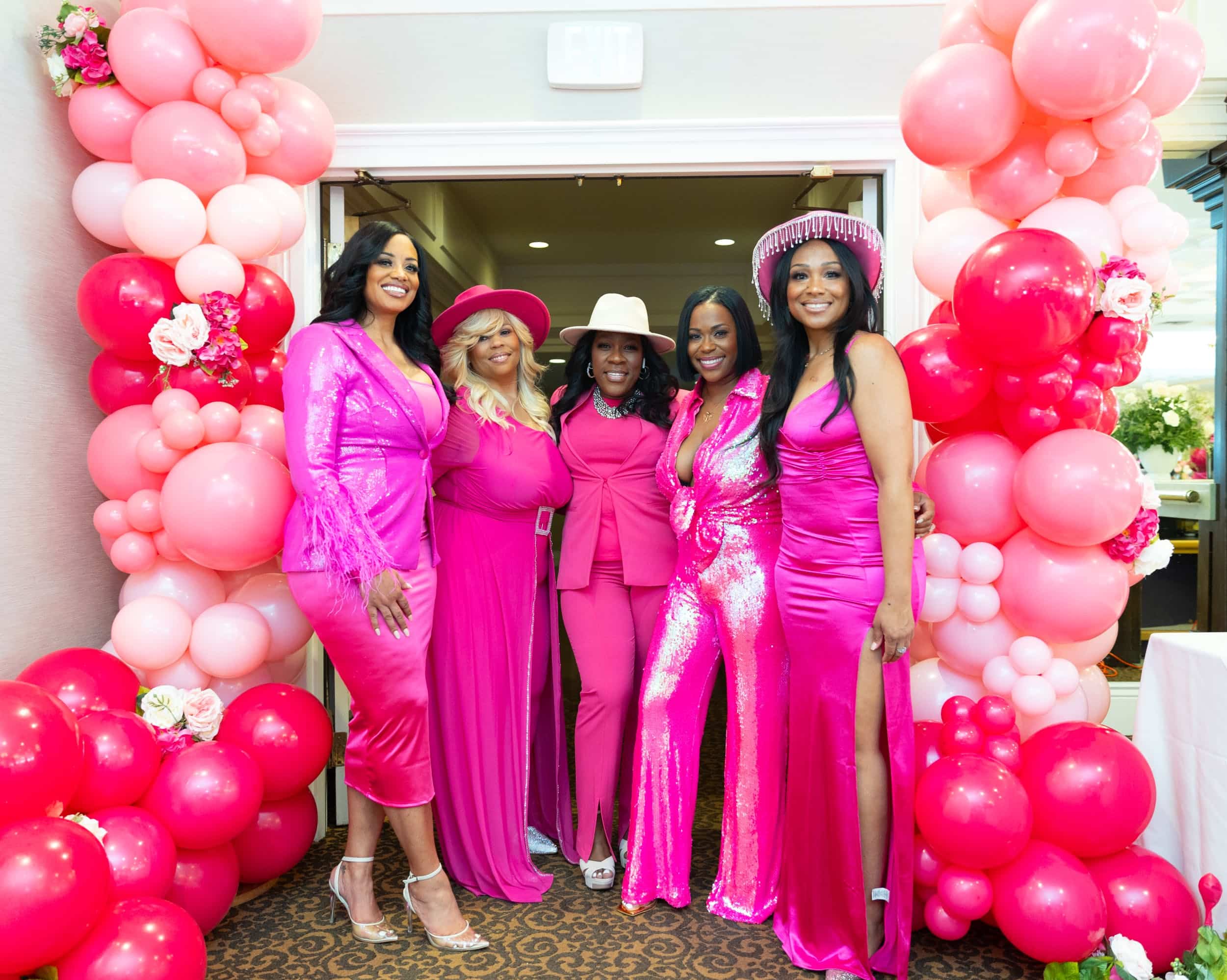 Group of women in pink attire posing at pink balloon arch entryway