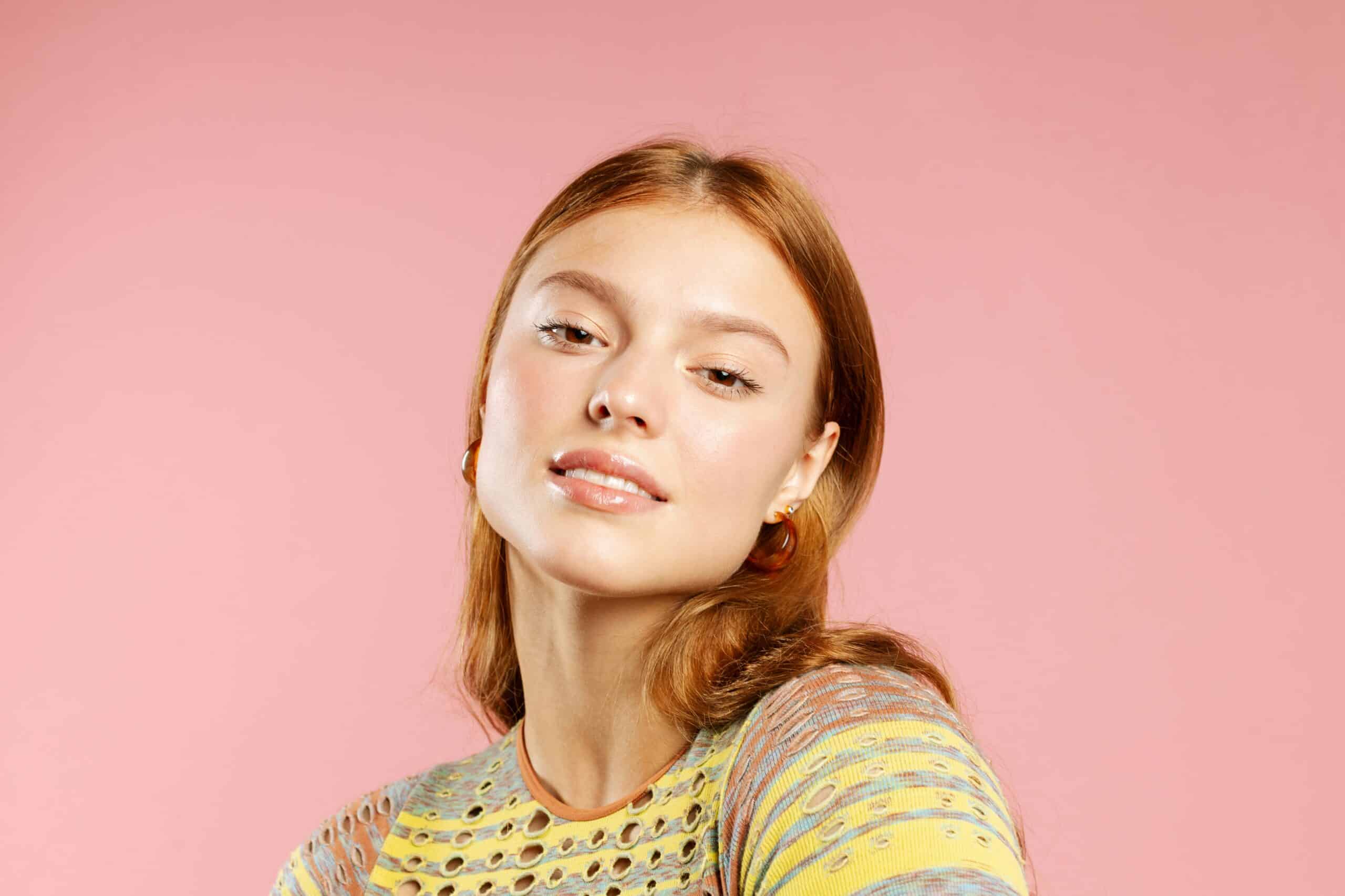 Woman with red hair posing with playful smirk against pink studio backdrop