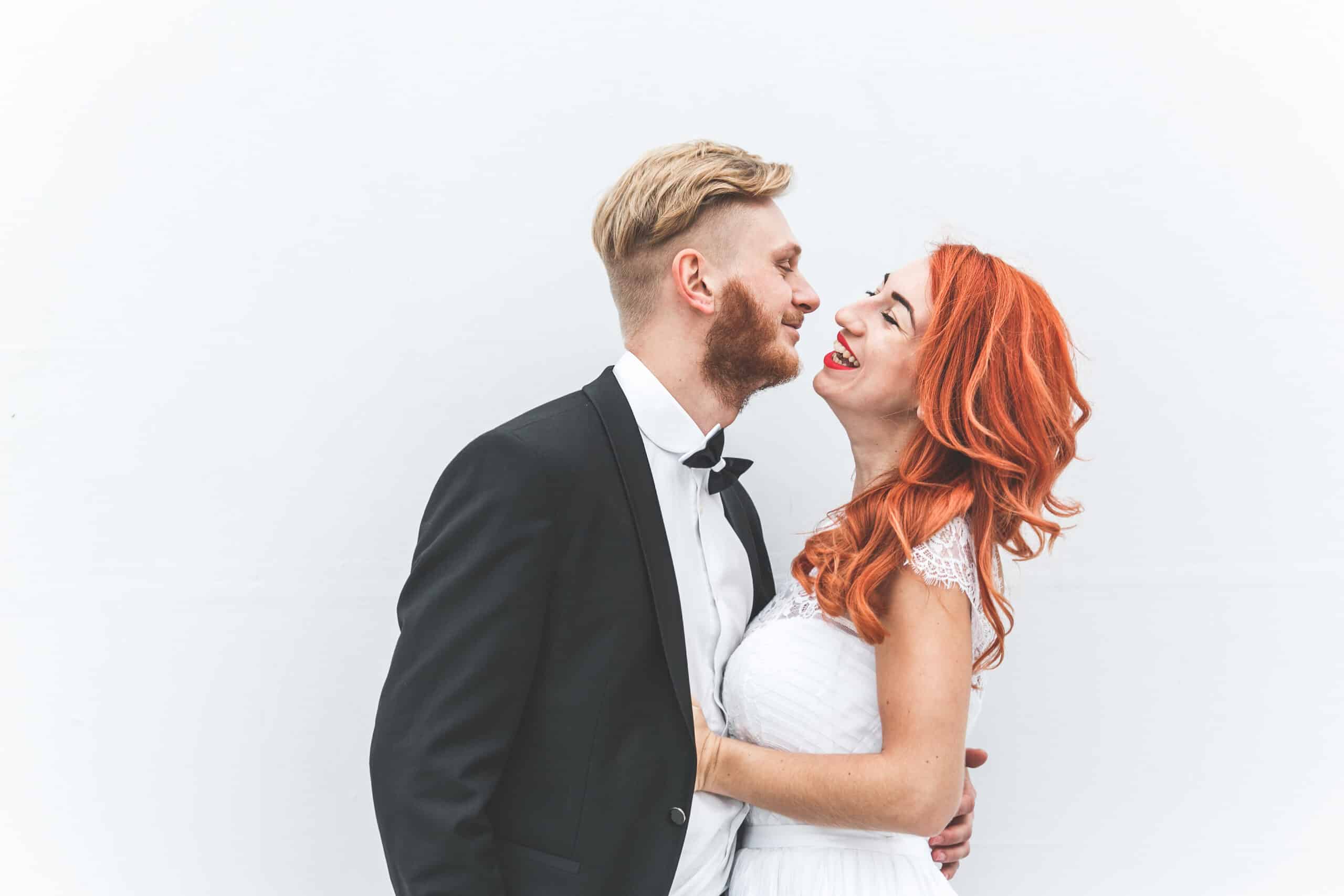 Bride with red hair and groom kissing playfully at white wall backdrop