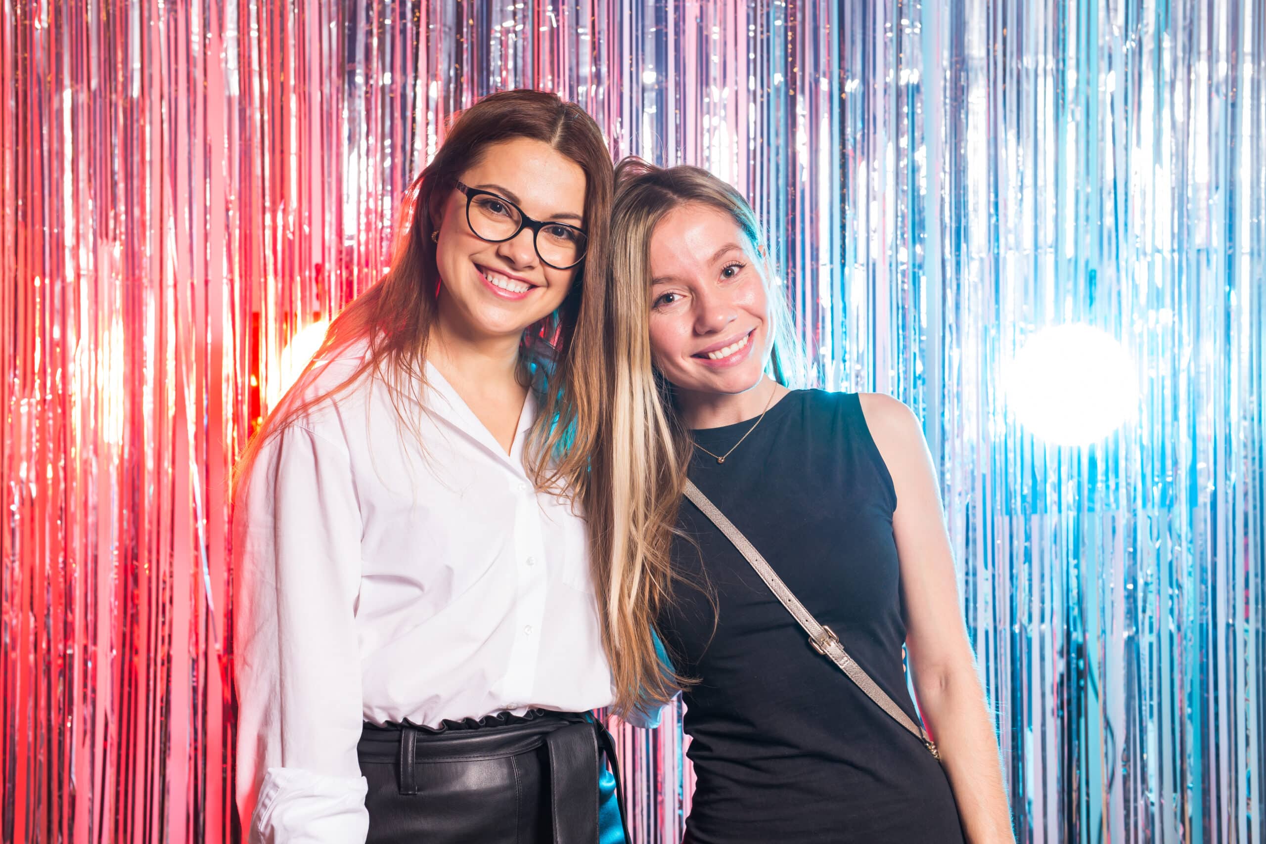 Two women smiling closely together at silver tinsel curtain backdrop