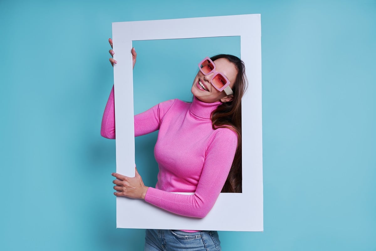 Young woman in pink turtleneck posing through white picture frame prop