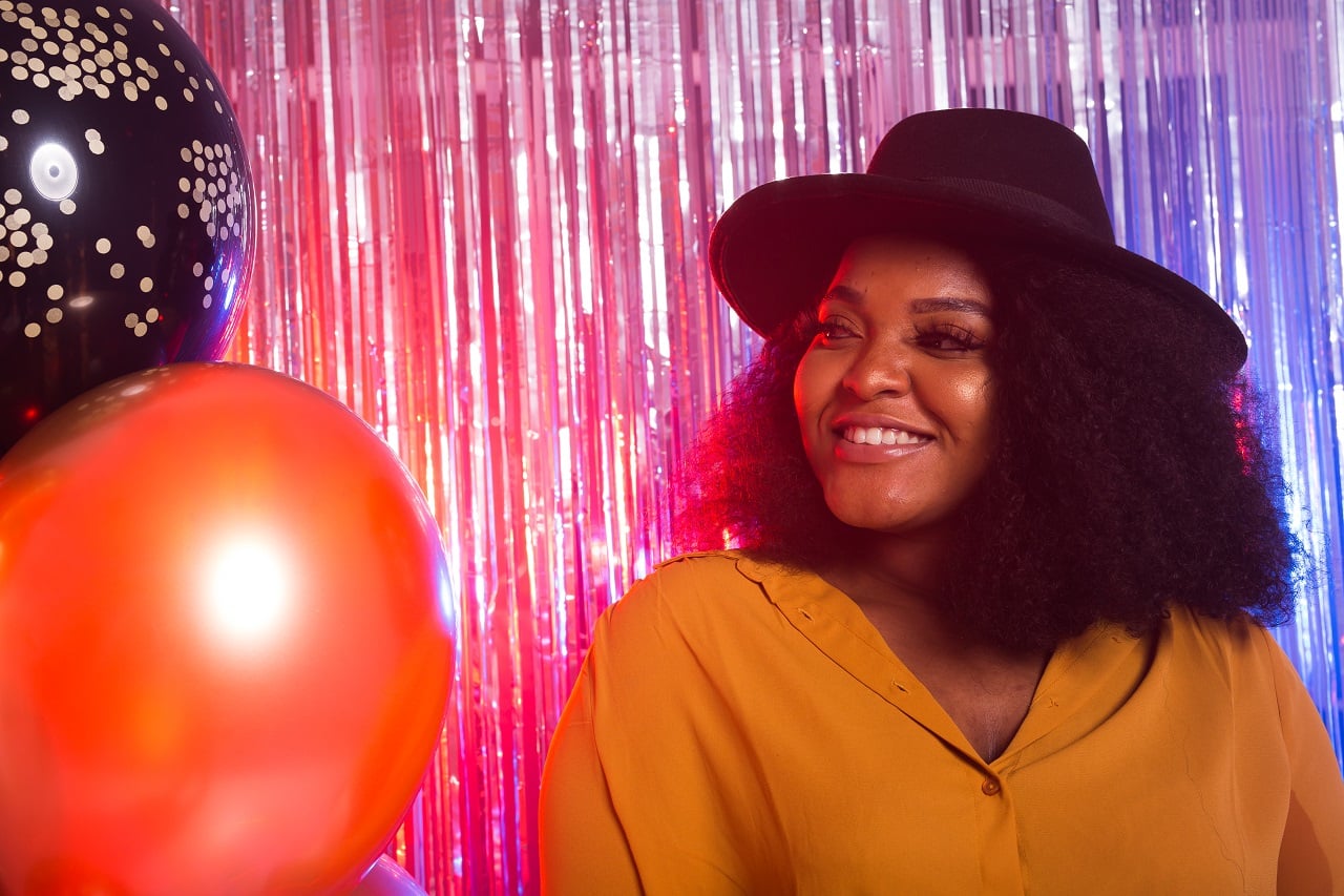 Woman in fedora hat smiling at colorful tinsel and balloon party backdrop