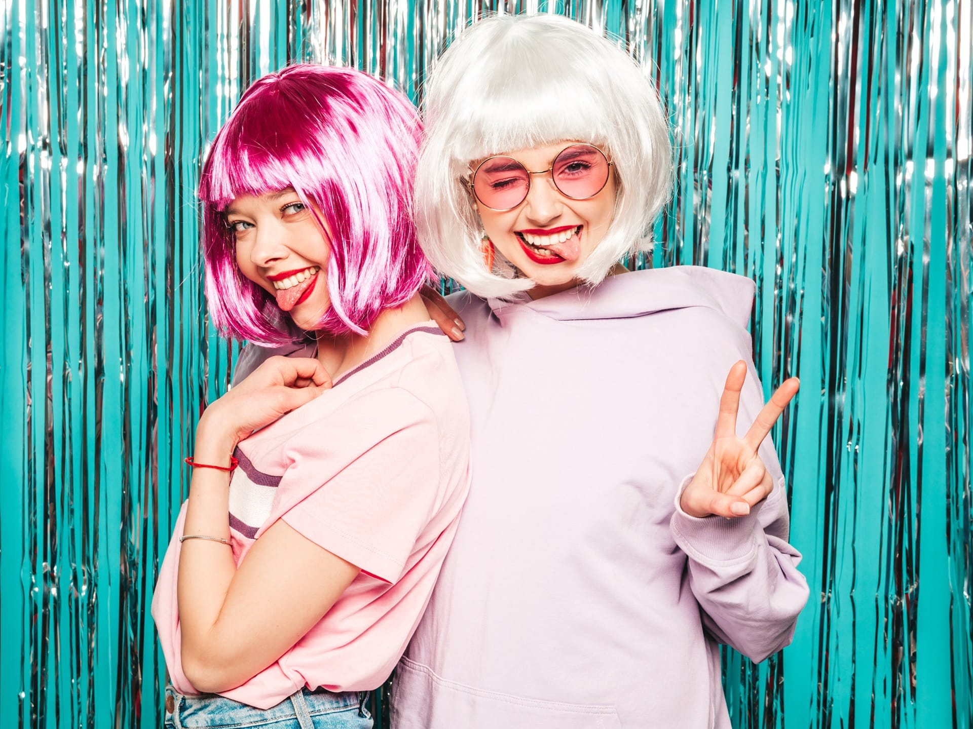 Two women in pink and white wigs posing at teal tinsel curtain backdrop
