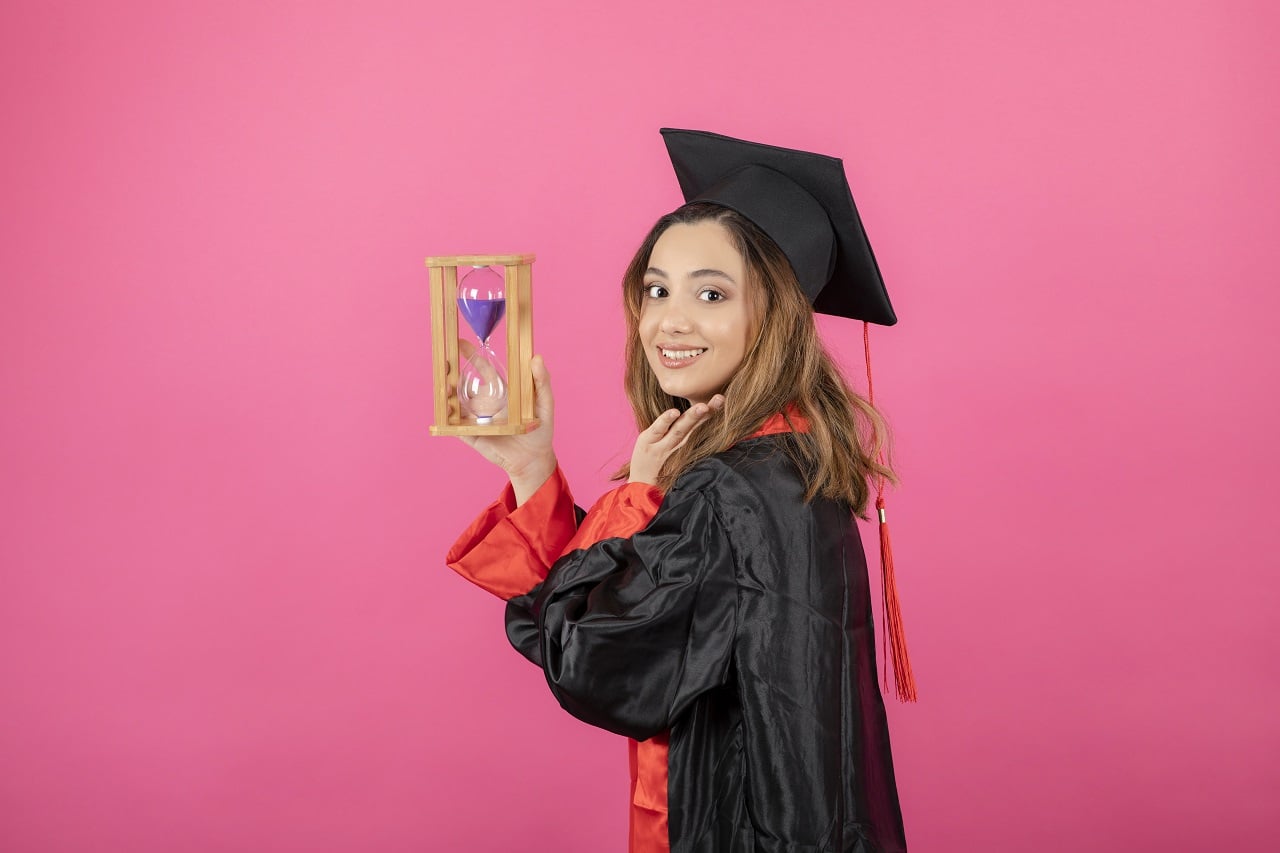 Female graduate in cap and gown holding hourglass at pink studio backdrop