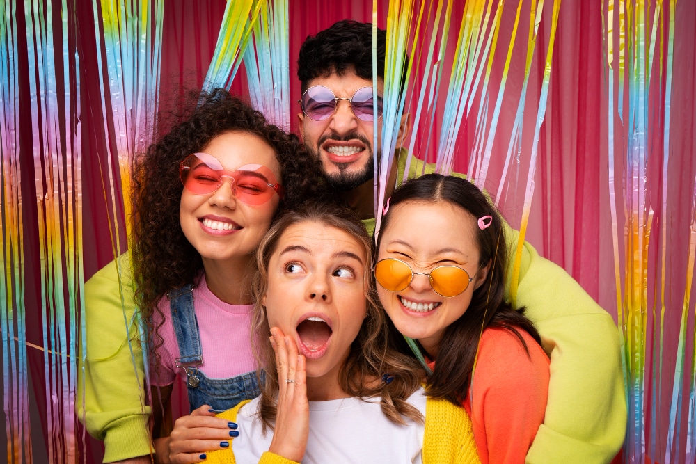 Group of friends in colorful outfits posing with sunglasses at rainbow tinsel backdrop