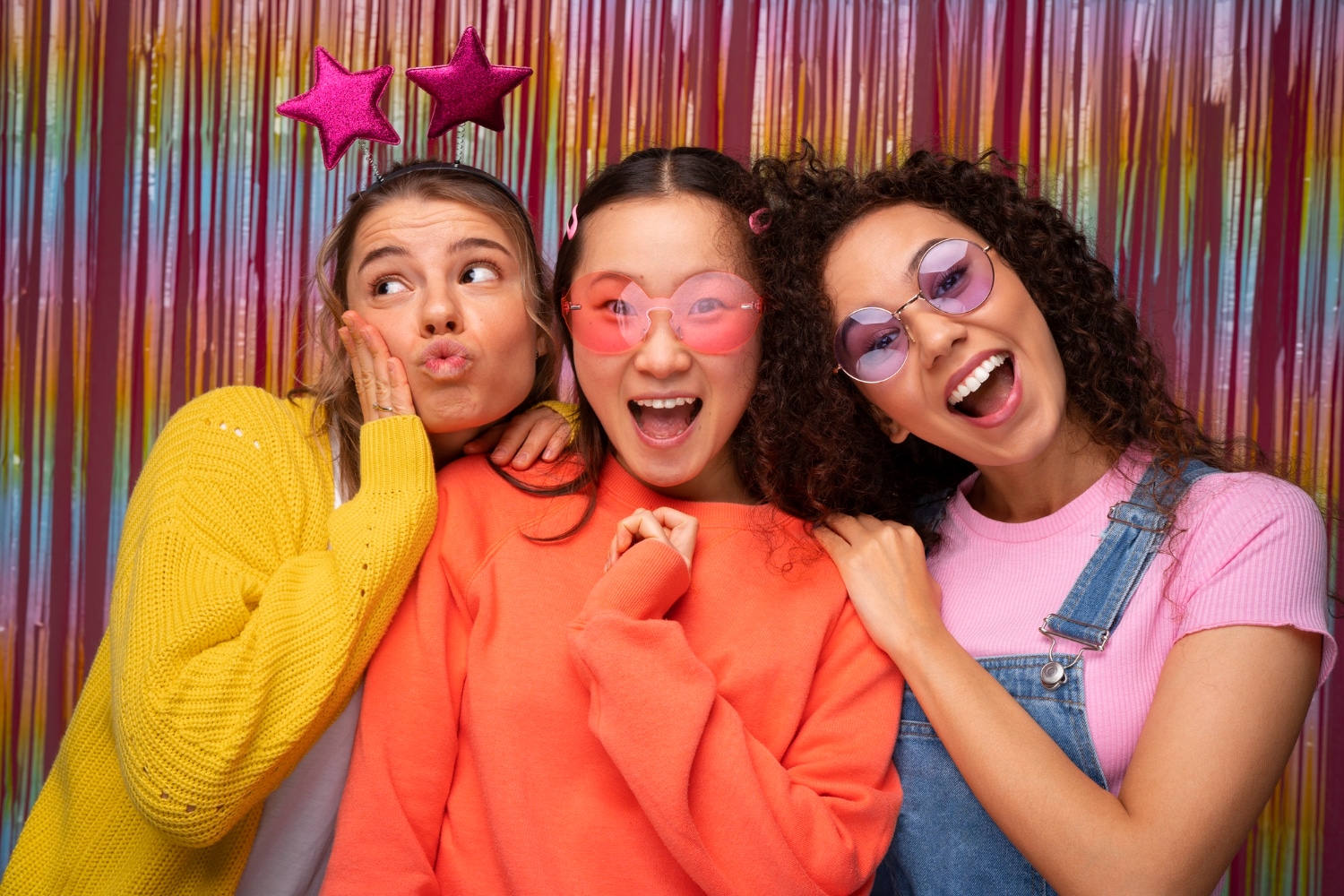 Three women in bold colors posing with star and heart sunglasses at pink backdrop