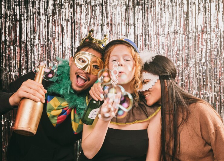 Three friends with carnival masks blowing bubbles at silver tinsel backdrop