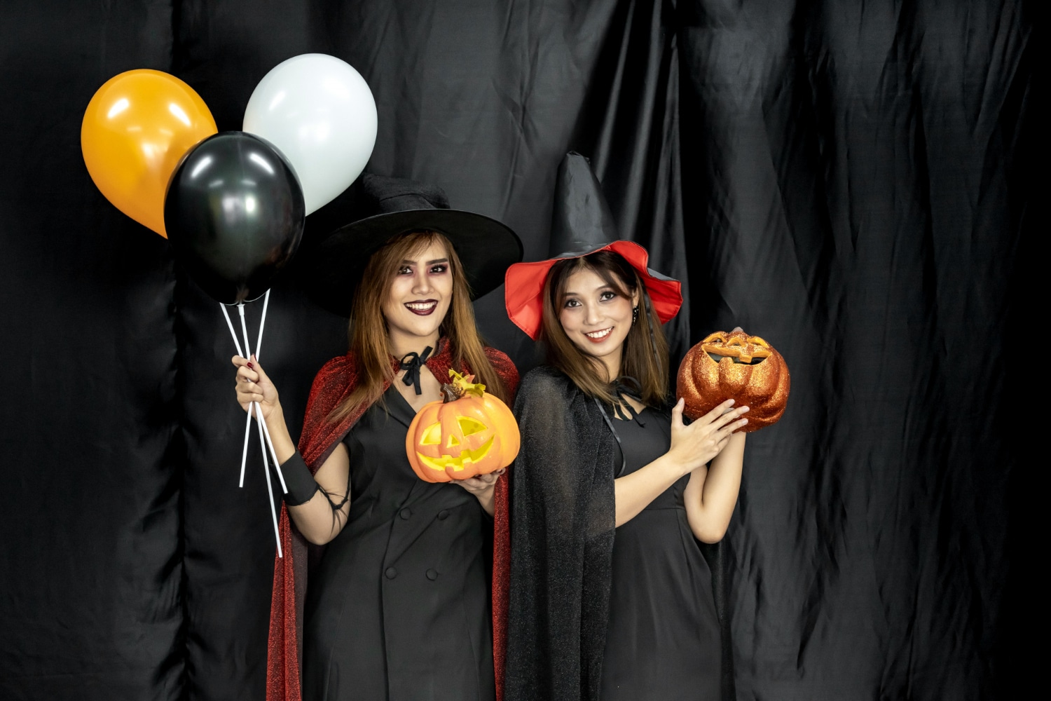 Two women in witch costumes holding pumpkins and balloons at black backdrop