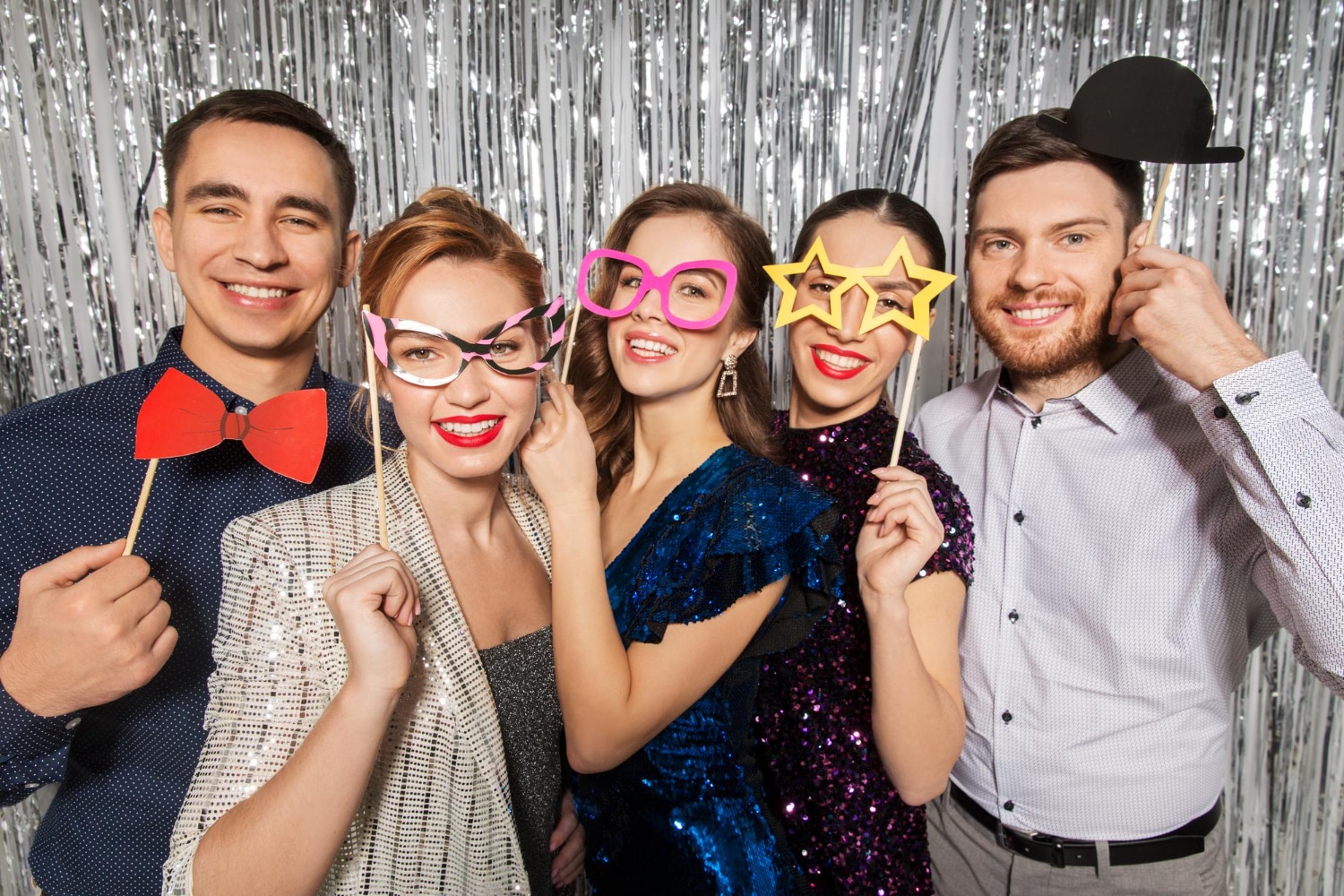 Group of five friends posing with assorted colorful photo booth props at silver backdrop