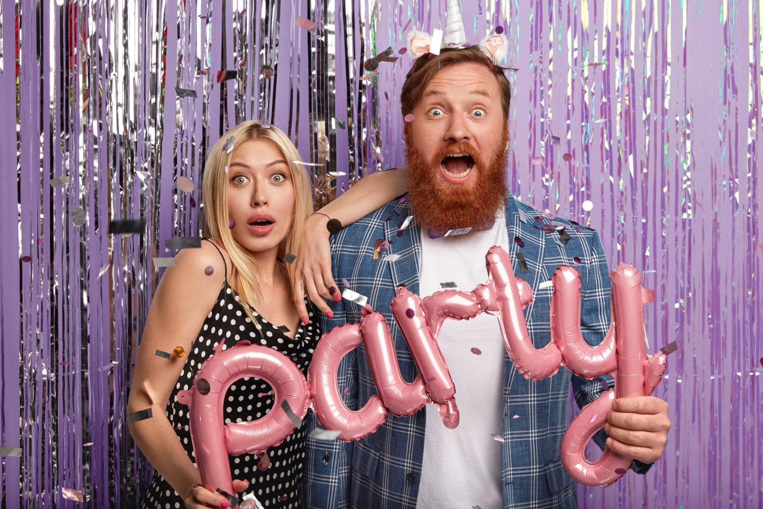 Couple holding pink Party balloons making surprised faces at purple tinsel backdrop