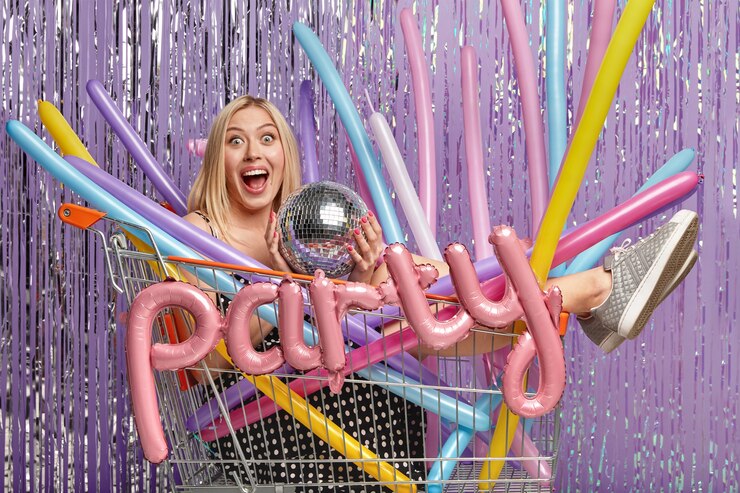 Blonde woman in shopping cart holding pink Party balloons at purple tinsel backdrop