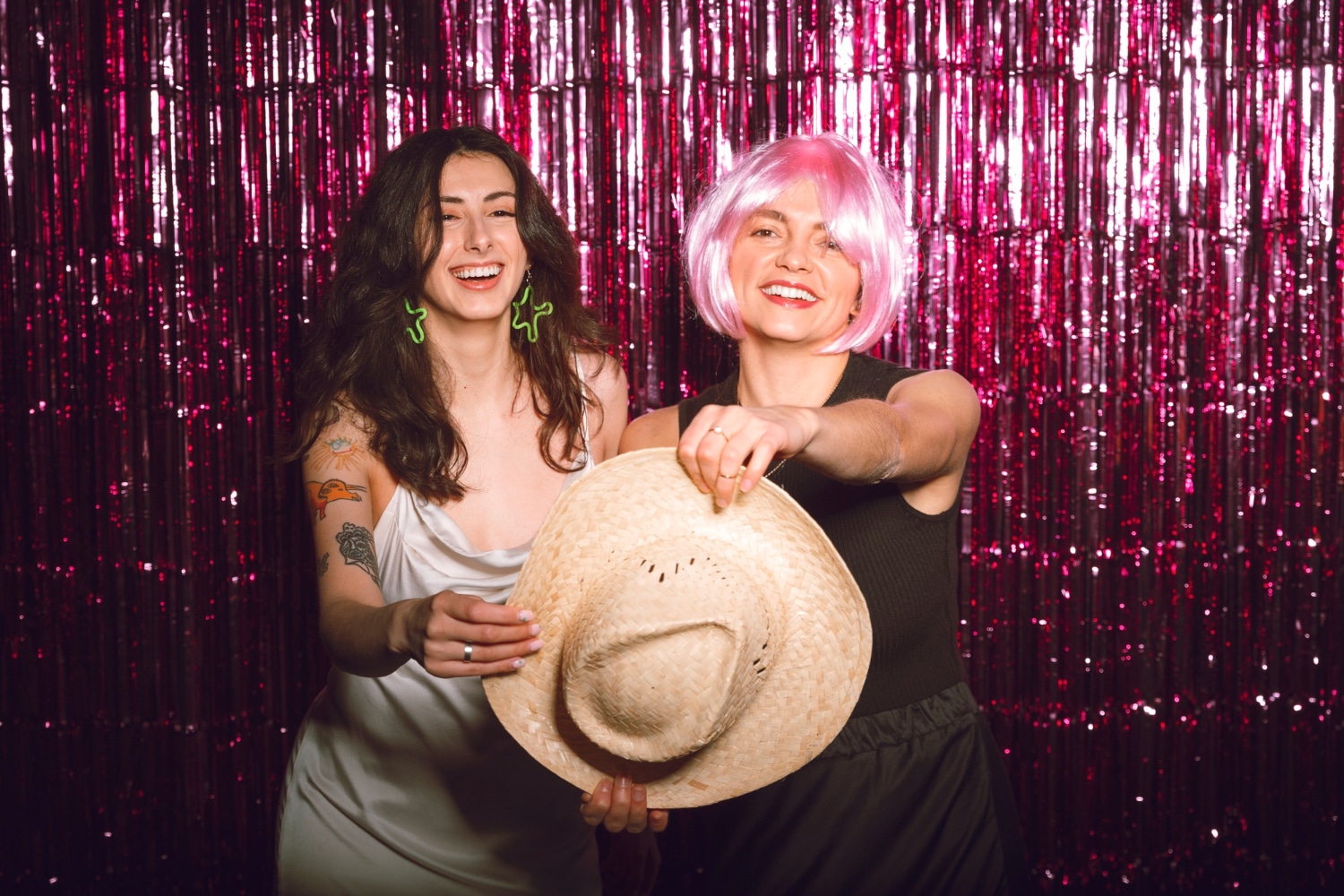 Two women in white outfits with cowboy hat and pink wig at hot pink tinsel backdrop