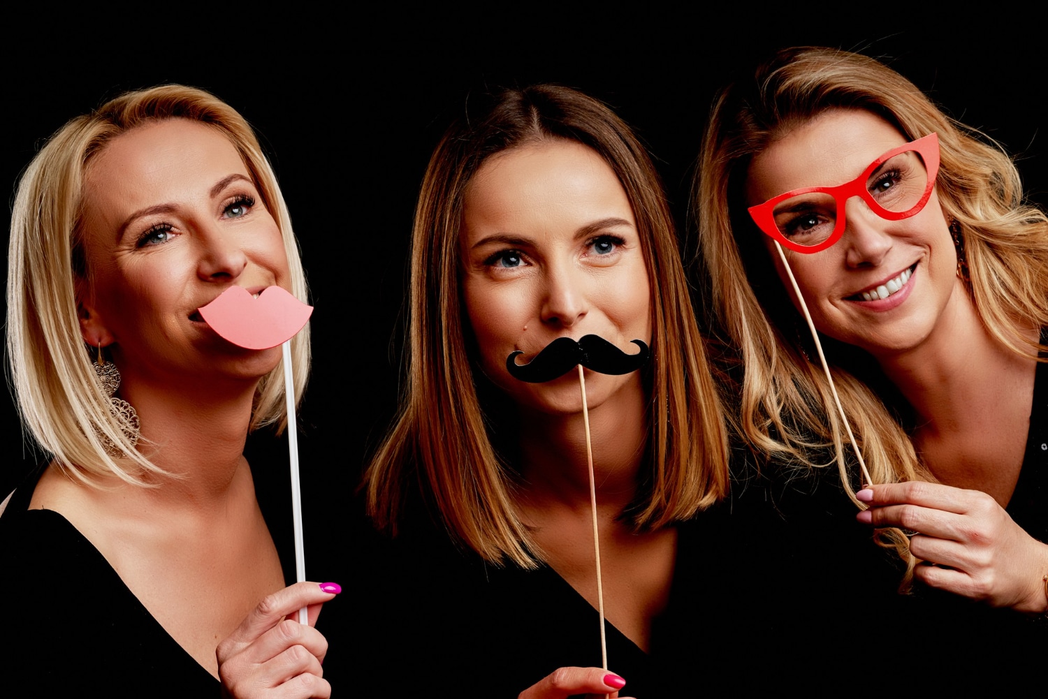 Three women holding lip and mustache props at black photo booth backdrop