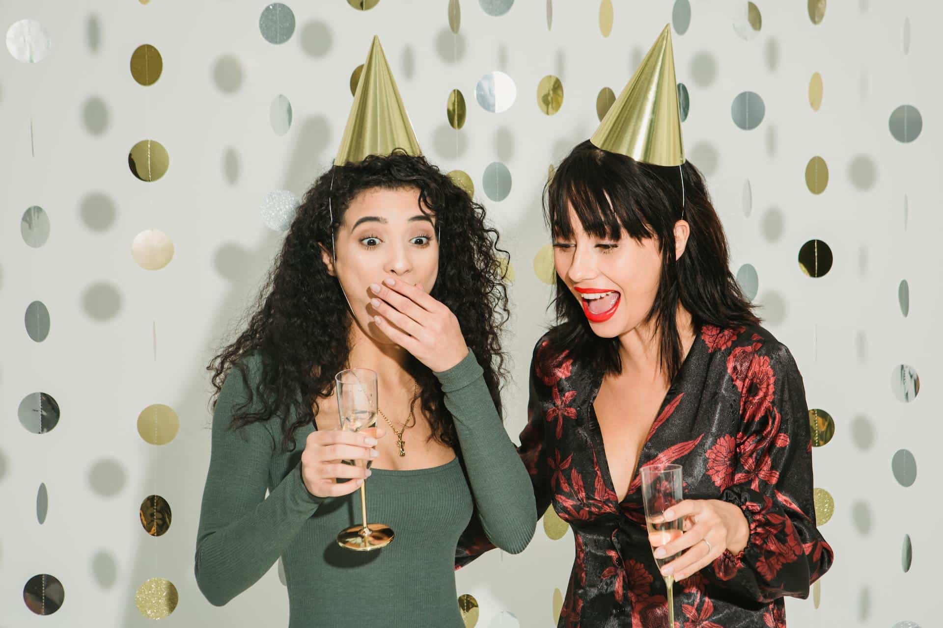 Two women in party hats celebrating with champagne at gold confetti backdrop
